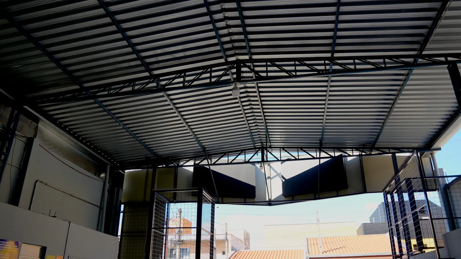 Vista interna de barracão com estrutura metálica escura e telhamento, aberto para a rua
