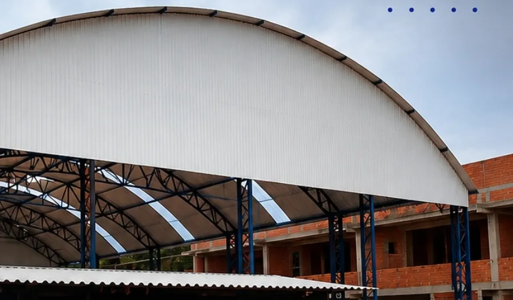 Cobertura metálica em arco com estrutura treliçada e pilares azuis, sobre construção em alvenaria